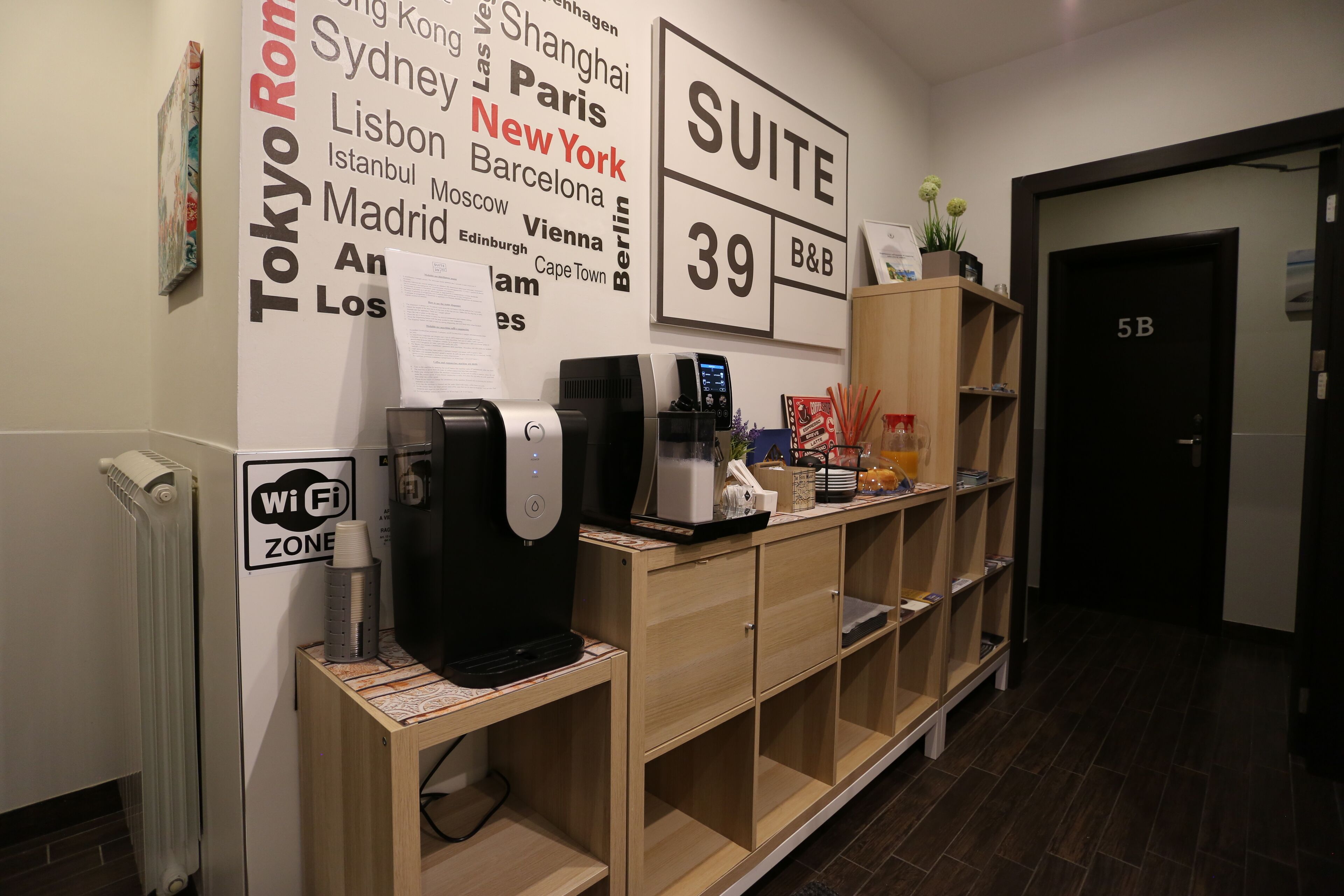Zona de recepción de hotel con un cartel que enumera varias ciudades, un letrero de 'SUITE 39', un letrero de 'Wi Fi ZONE' y una máquina de café.