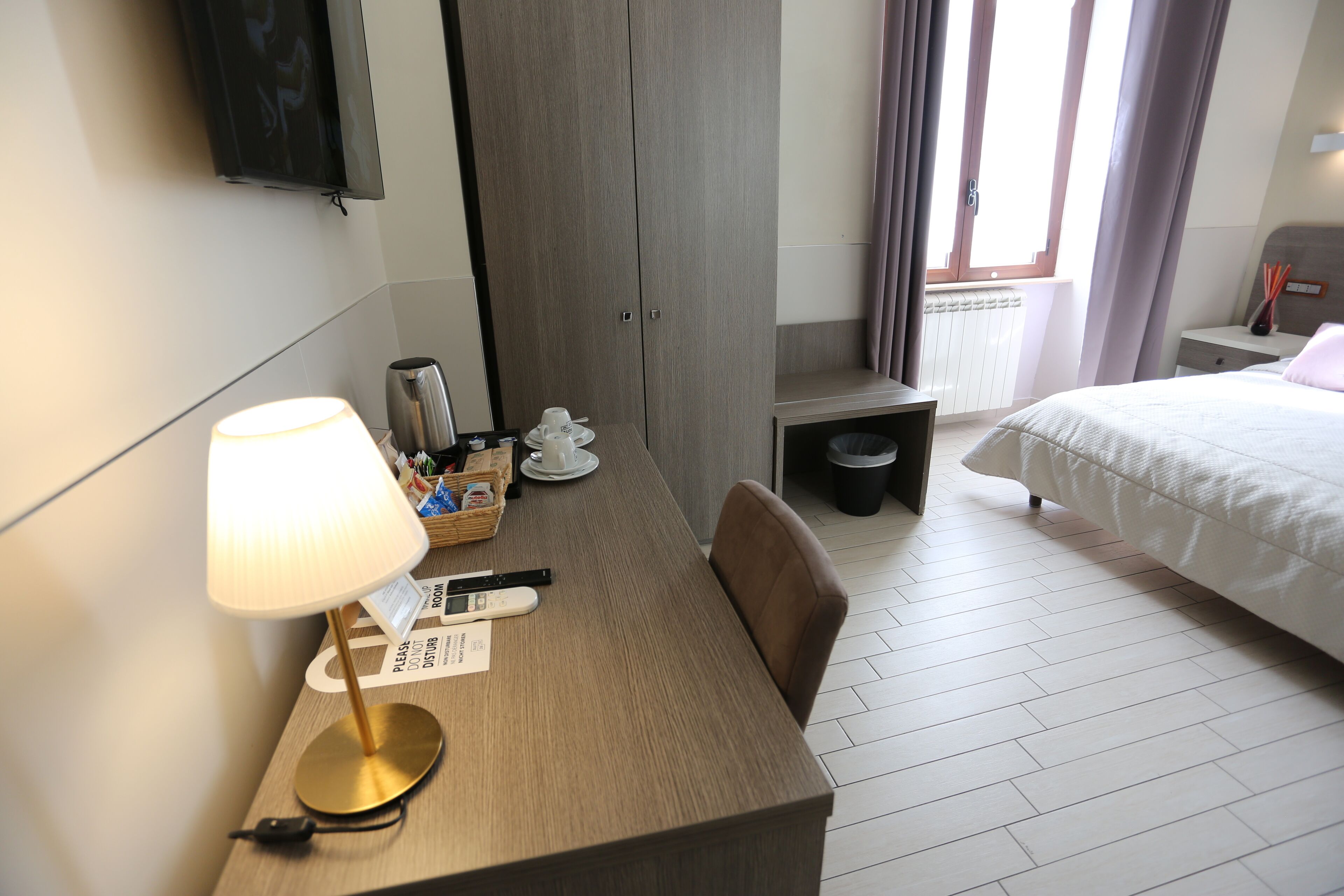 A hotel room with a wooden desk, a lamp, a bed, a wardrobe, and a window with curtains.