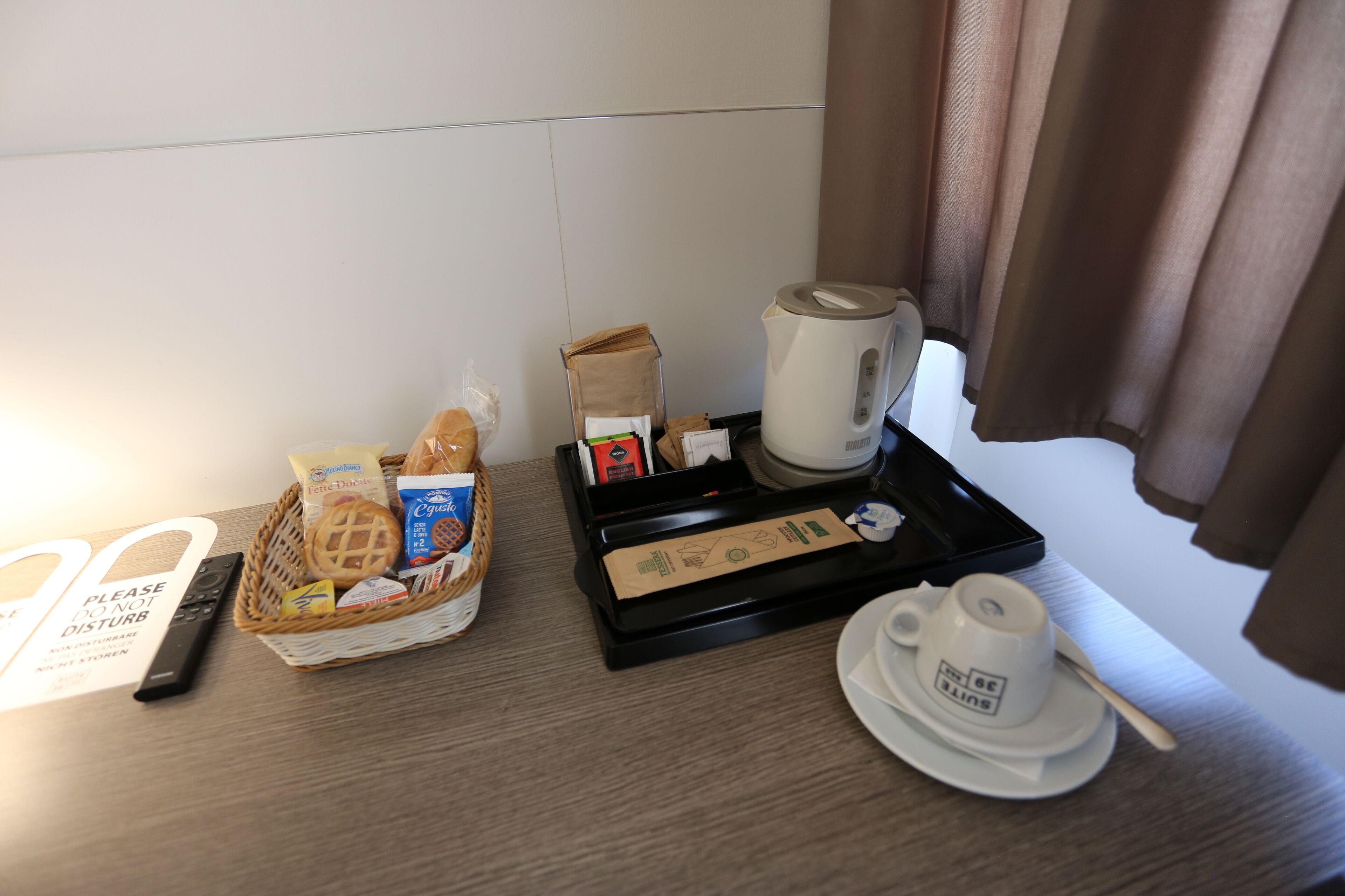 Et hotelværelse med bakke til morgenmad, en kurv med brød, en kaffemaskine samt kop og underkop.