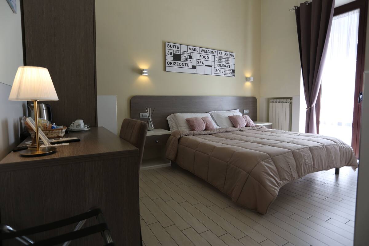 A hotel room with a bed, a desk with a lamp, a wall-mounted sign, and a window with curtains.