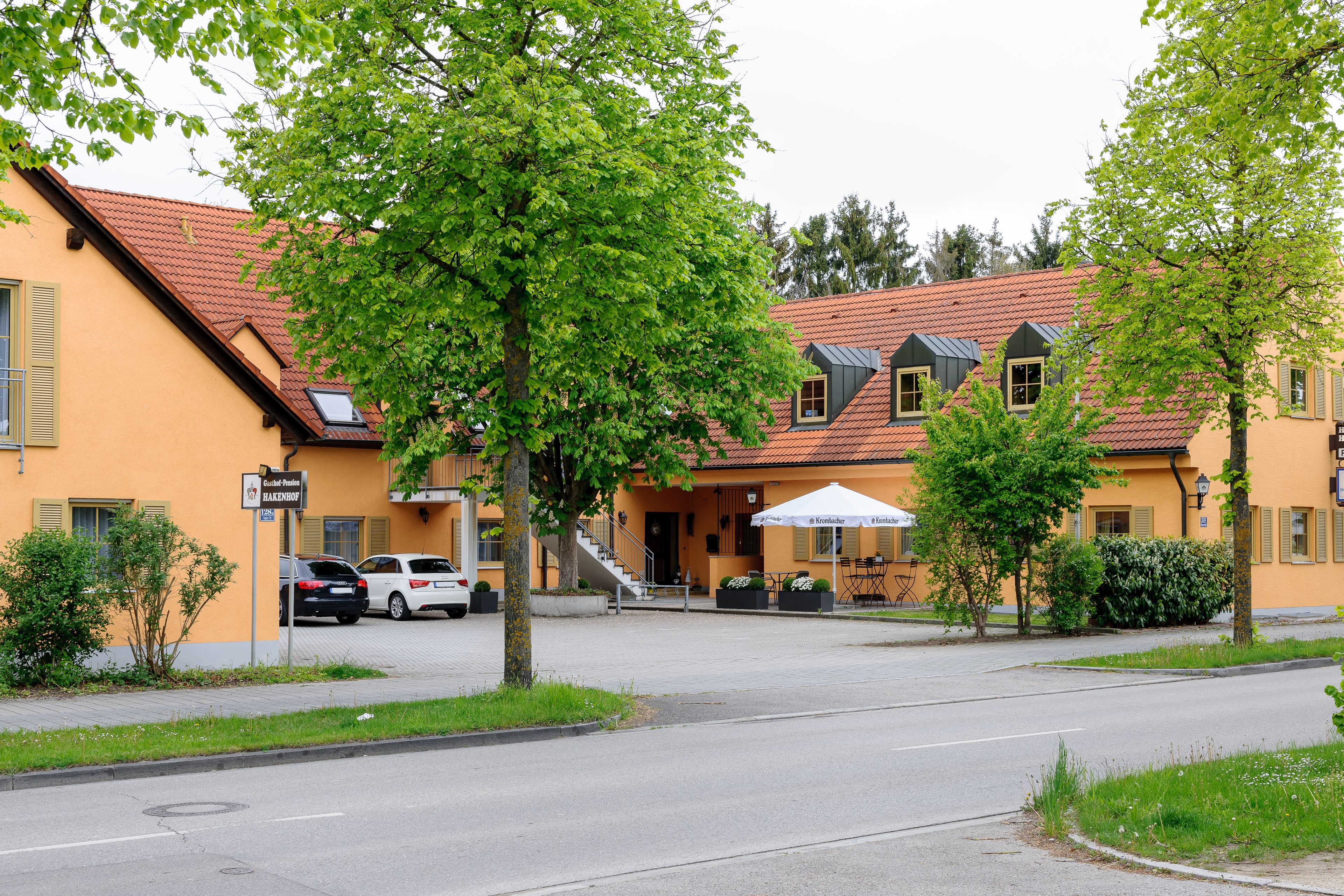 En bygning med røde tegltagsten, en gul facade og et skilt, der angiver en restaurant. Der er biler parkeret foran, og et træ i forgrunden.