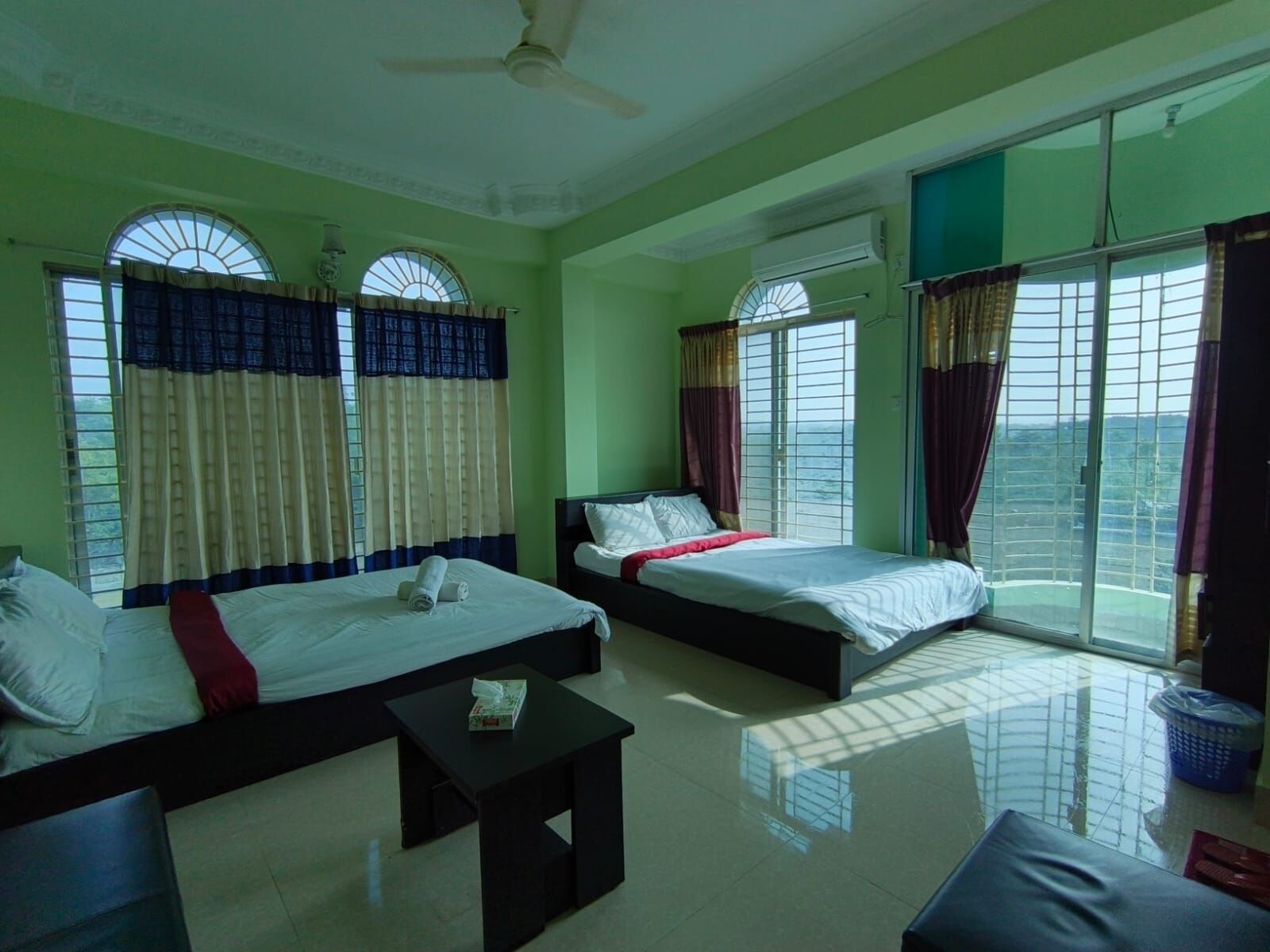 Habitación de hotel con dos camas, ventilador de techo, ventanas grandes con cortinas, una mesita pequeña y vistas al exterior.
