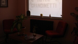 A dimly lit room with a large window displaying the text "BENVENUTI HOTEL GRANOINETTI". There is a potted plant, a small table with a vase, and two chairs. - Grandinetti Hotel (Parenti)
