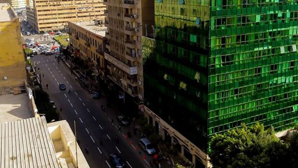 Un edificio de varios pisos con fachada de vidrio moderna, junto a una calle concurrida con vehículos y peatones.