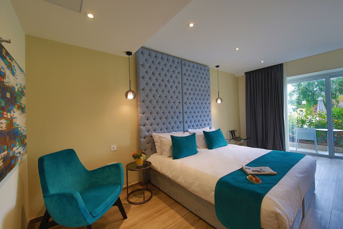 A bedroom with a large bed, a blue chair, a small table with a vase, and a view of the outdoors.