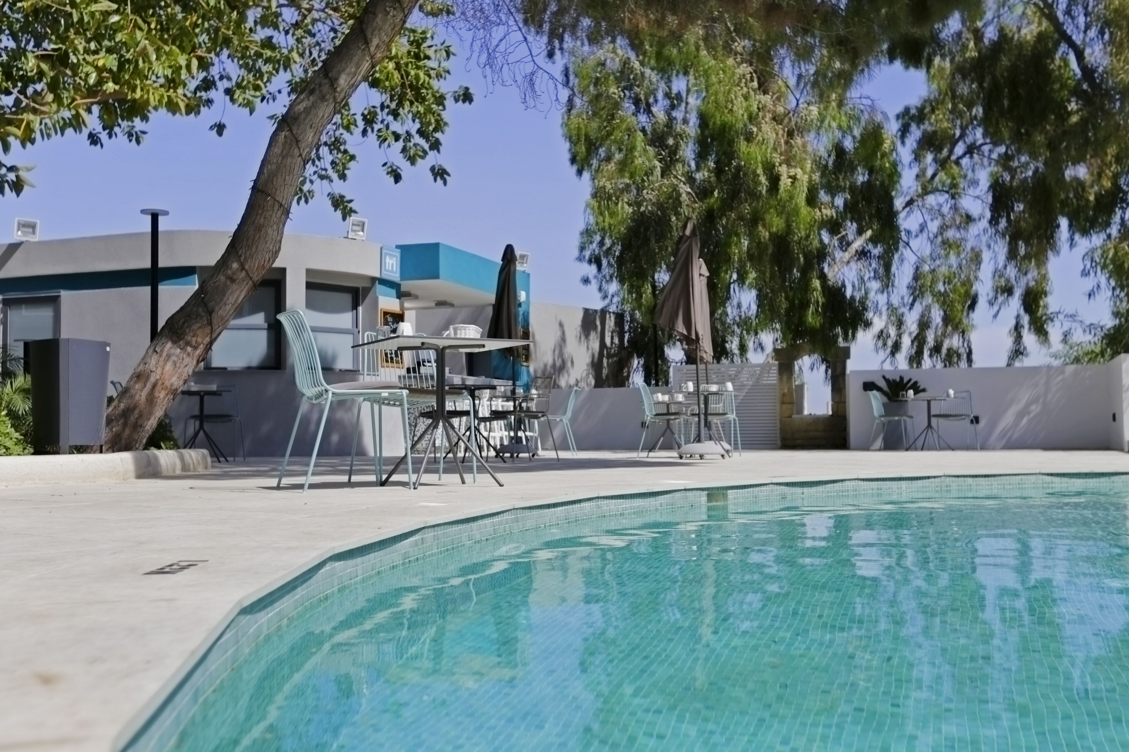 Una piscina amb gandules i taules, envoltada d'arbres i un edifici modern.