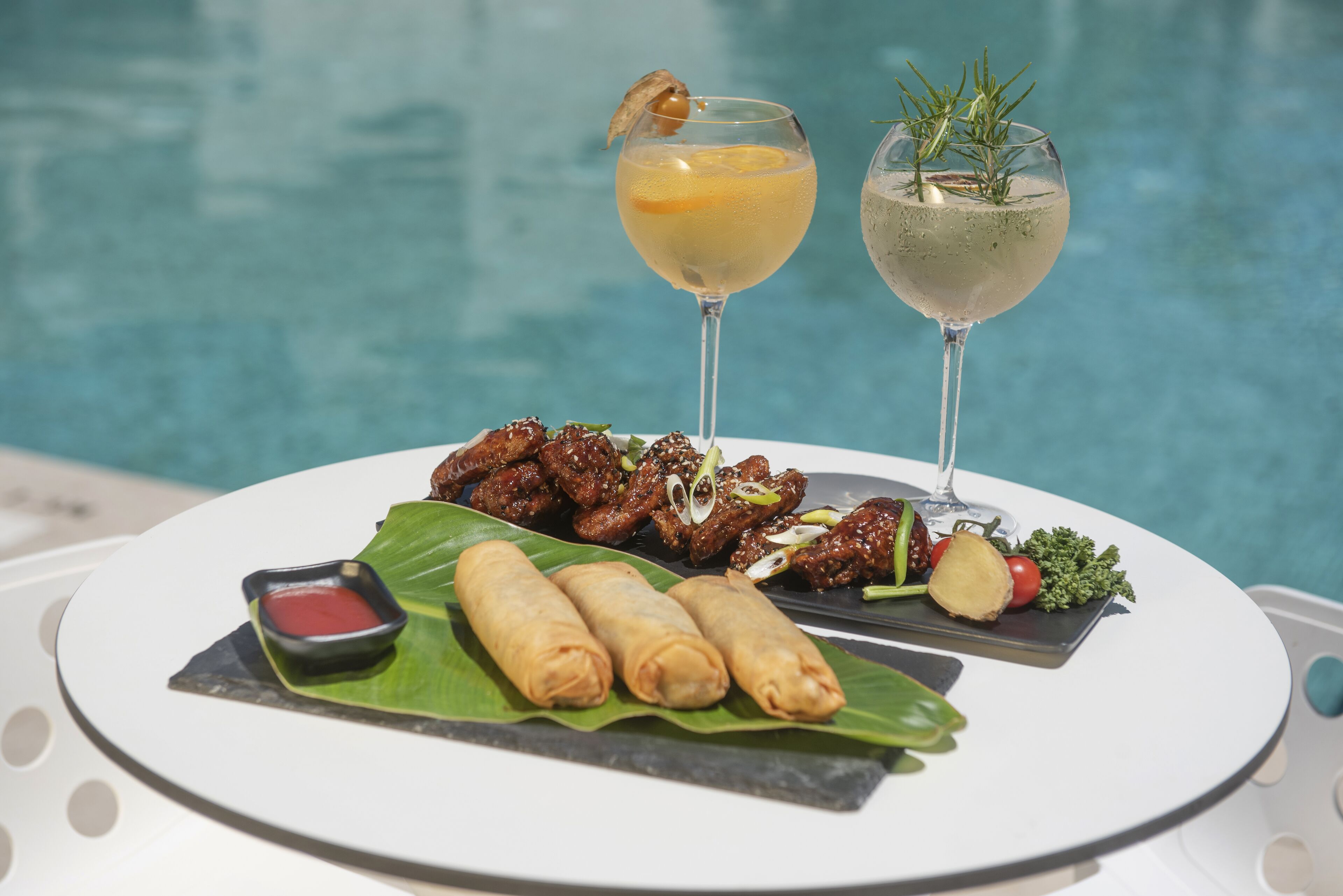 Une table à manger avec une assiette de nourriture, deux boissons et une piscine en arrière-plan.