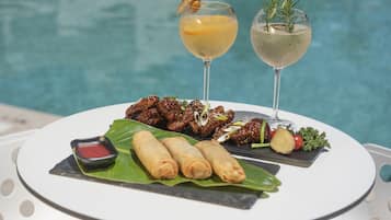 A dining table with a plate of food, two drinks, and a pool in the background.