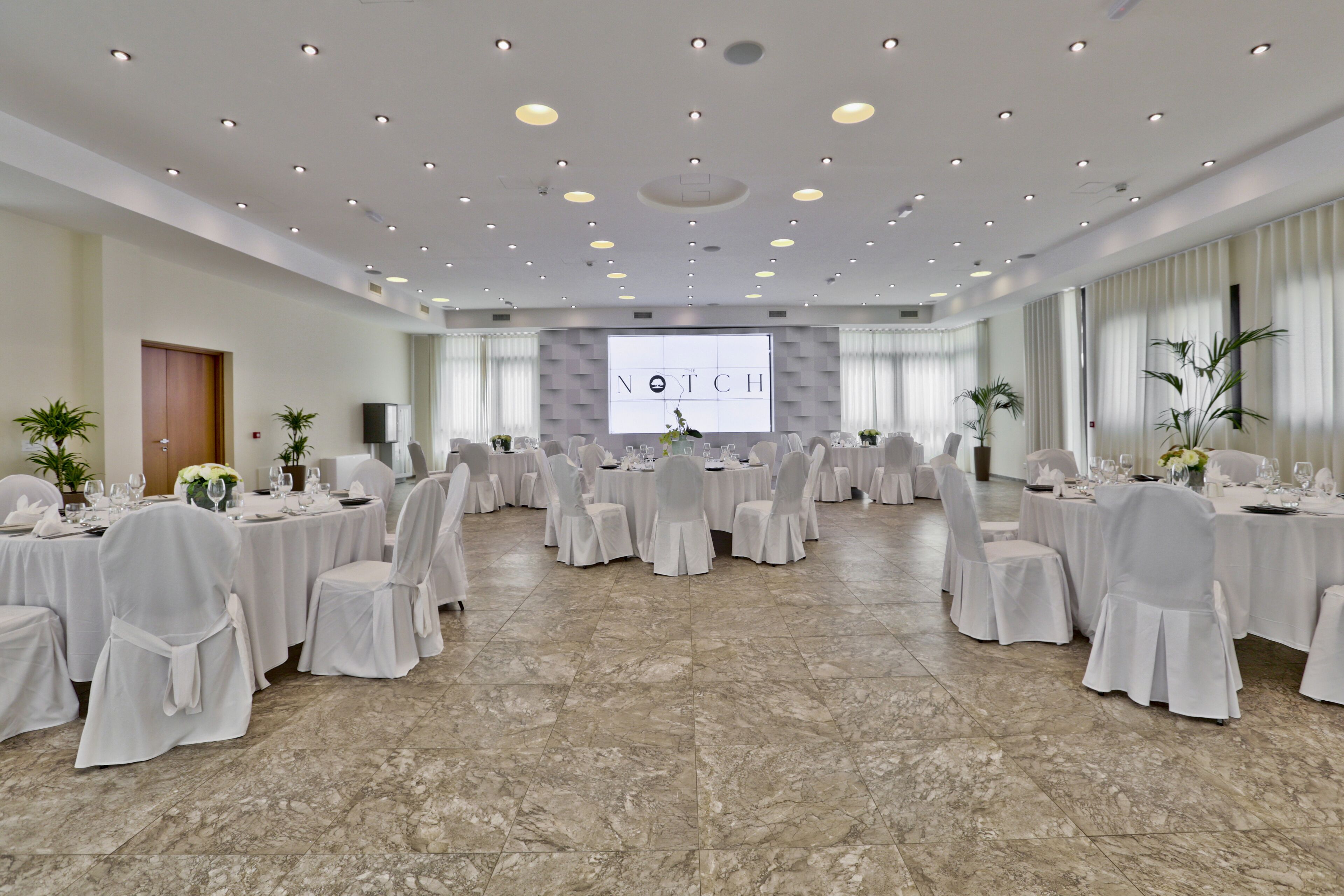 Uma sala de conferências com várias mesas redondas, toalhas brancas e cadeiras, uma tela grande na frente e piso de mármore.