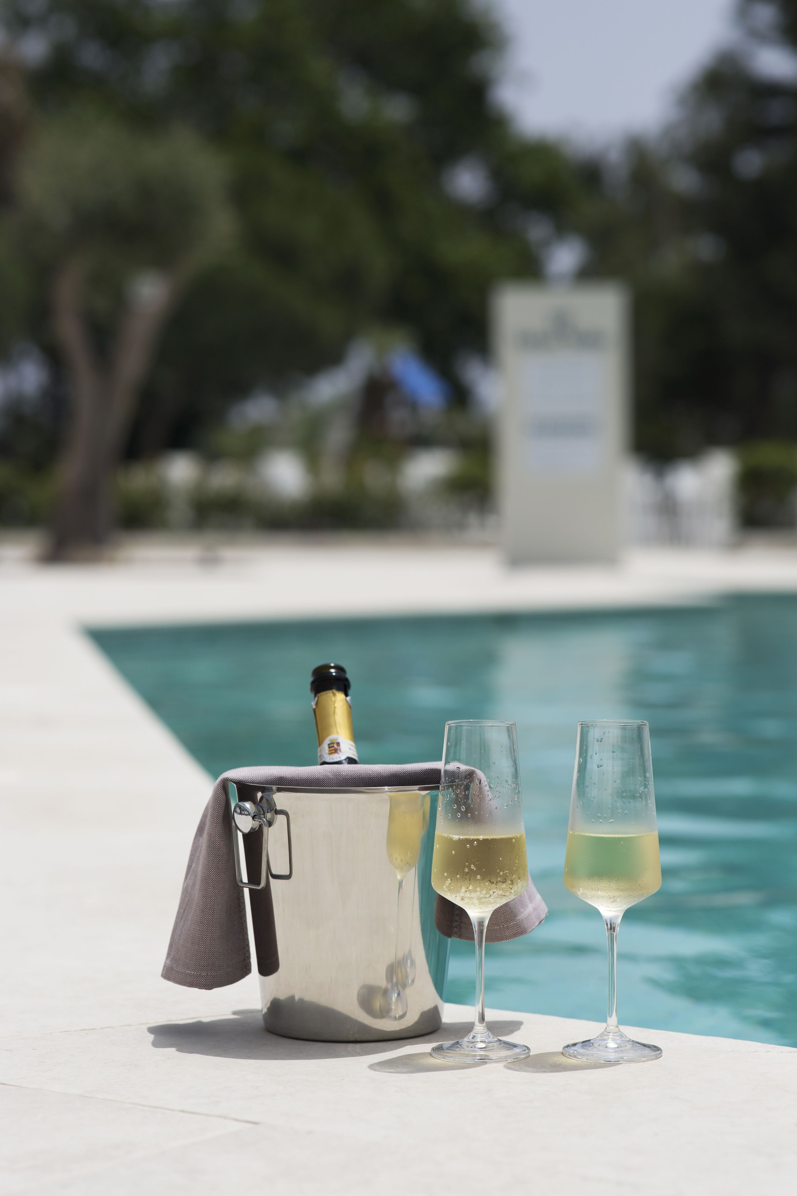 A champagne bottle and two glasses on a table by a pool with a white tiled edge.