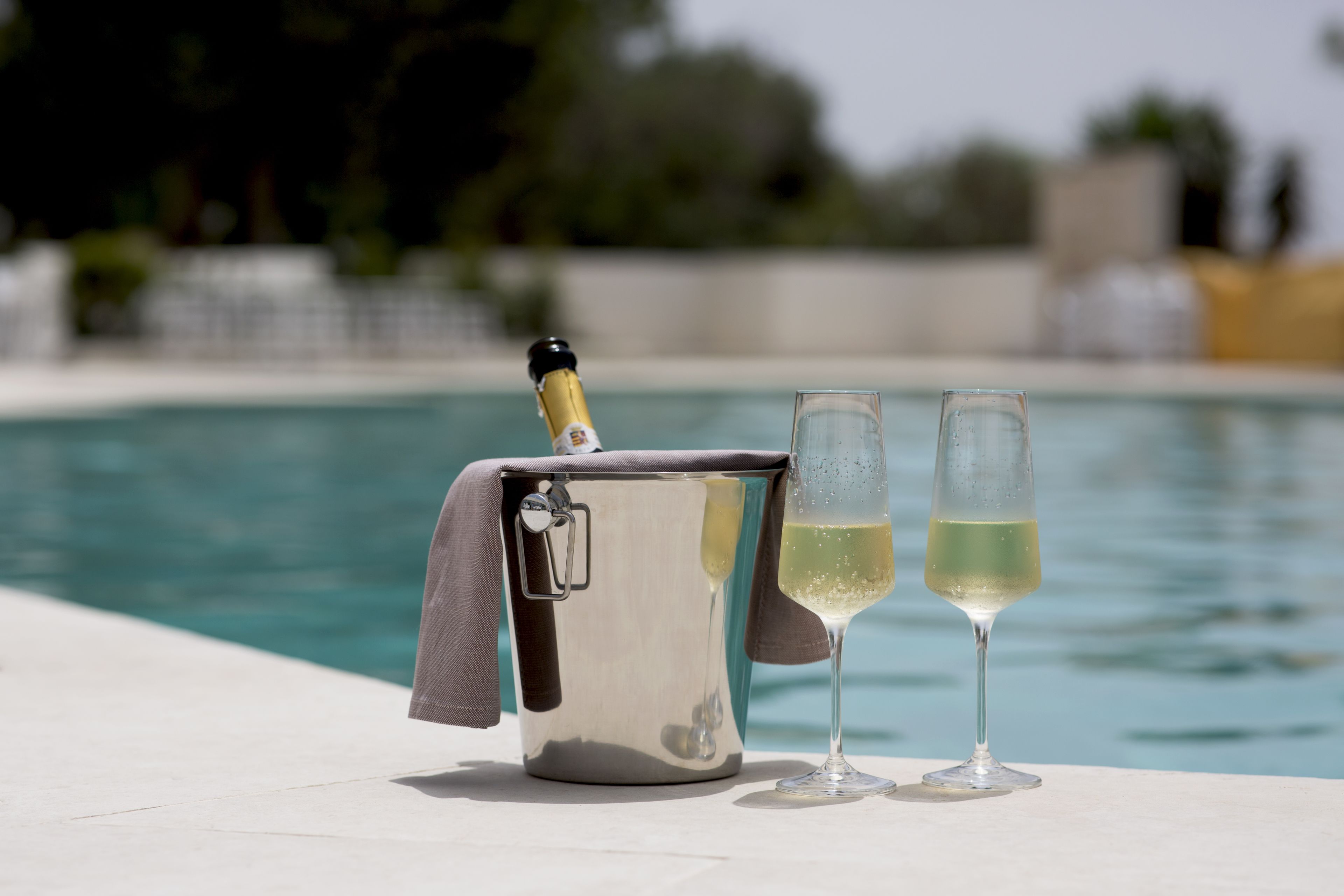 Un cubo metálico con un abrelatas y un paño, dos vasos con una bebida, y una piscina al fondo.
