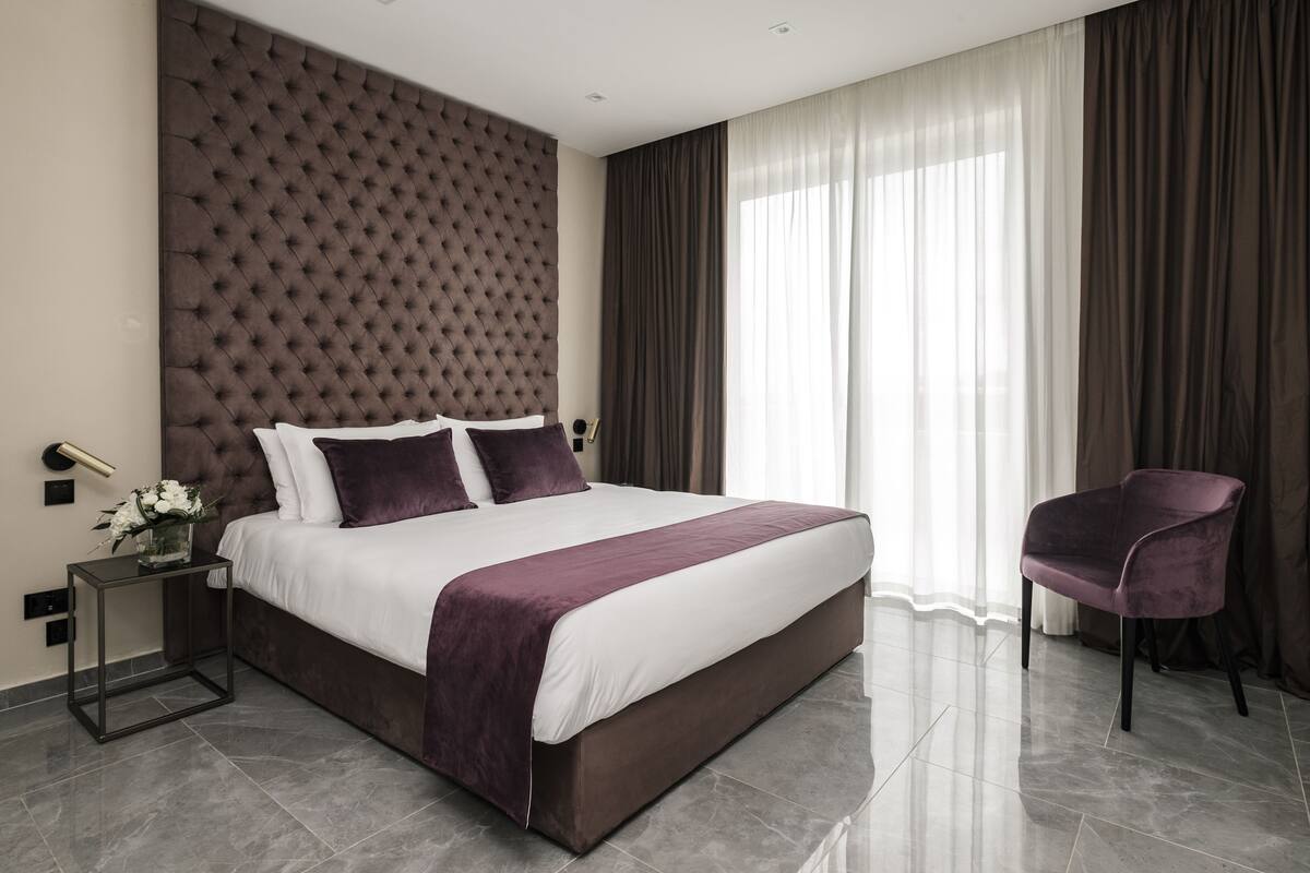 A modern bedroom with a tufted headboard, a large bed with white and purple bedding, a side table with a vase of flowers, and a chair.