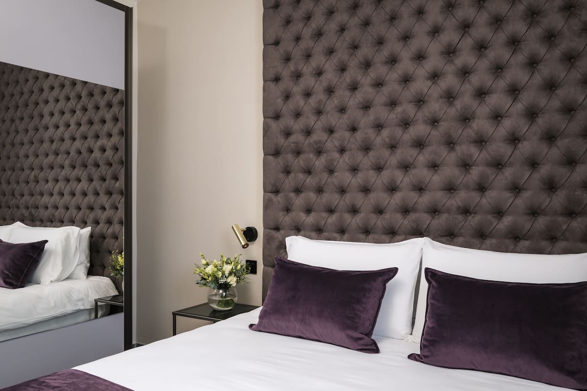 A modern hotel room with a tufted headboard, white bedding, and a small side table with a vase of flowers.