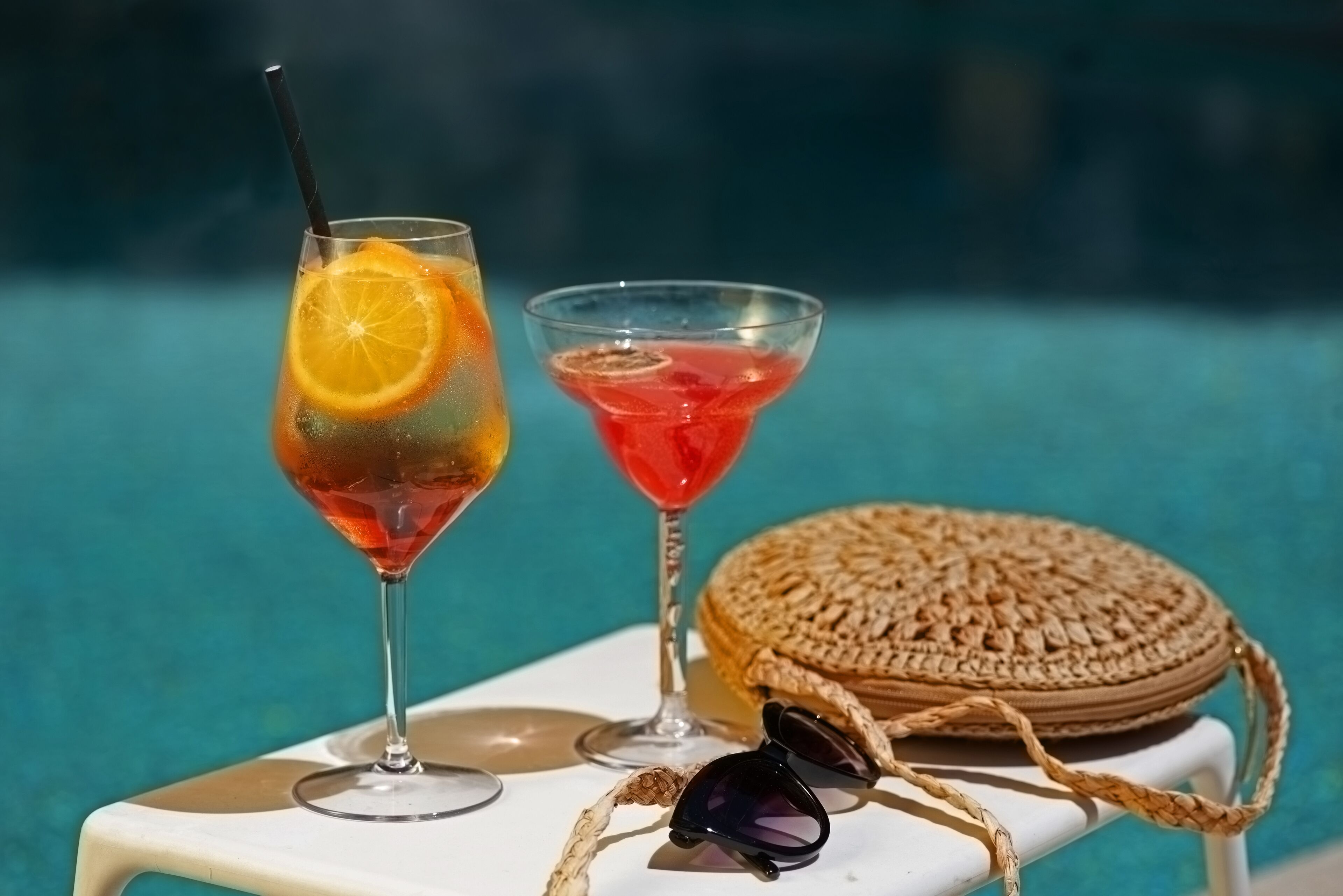 Dos bebidas sobre una mesa blanca junto a una piscina, con un bolso tejido y gafas de sol cerca.