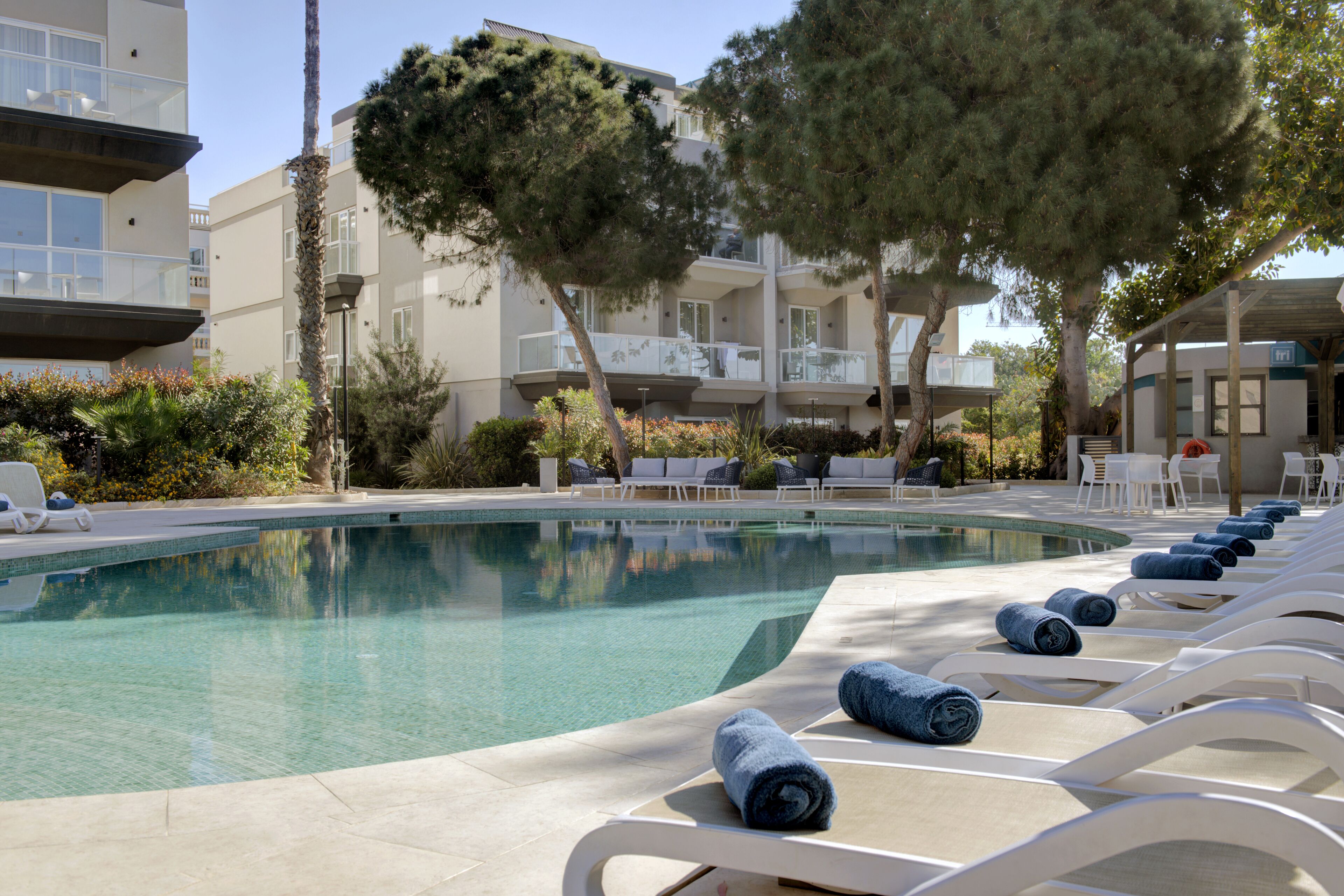 A pool area with lounge chairs and towels, surrounded by trees and buildings.