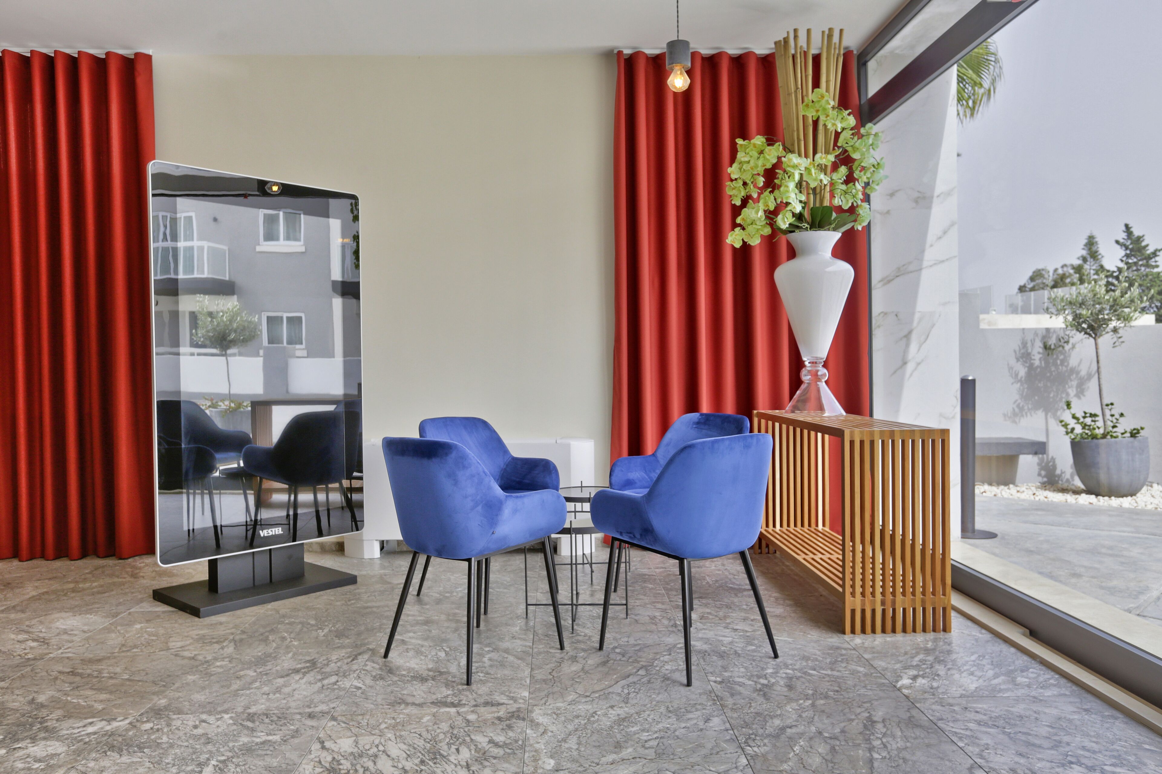 Una habitación de hotel moderna con un televisor de pantalla plana grande, dos sillas azules, una mesita pequeña, un mueble de madera y una planta en maceta.