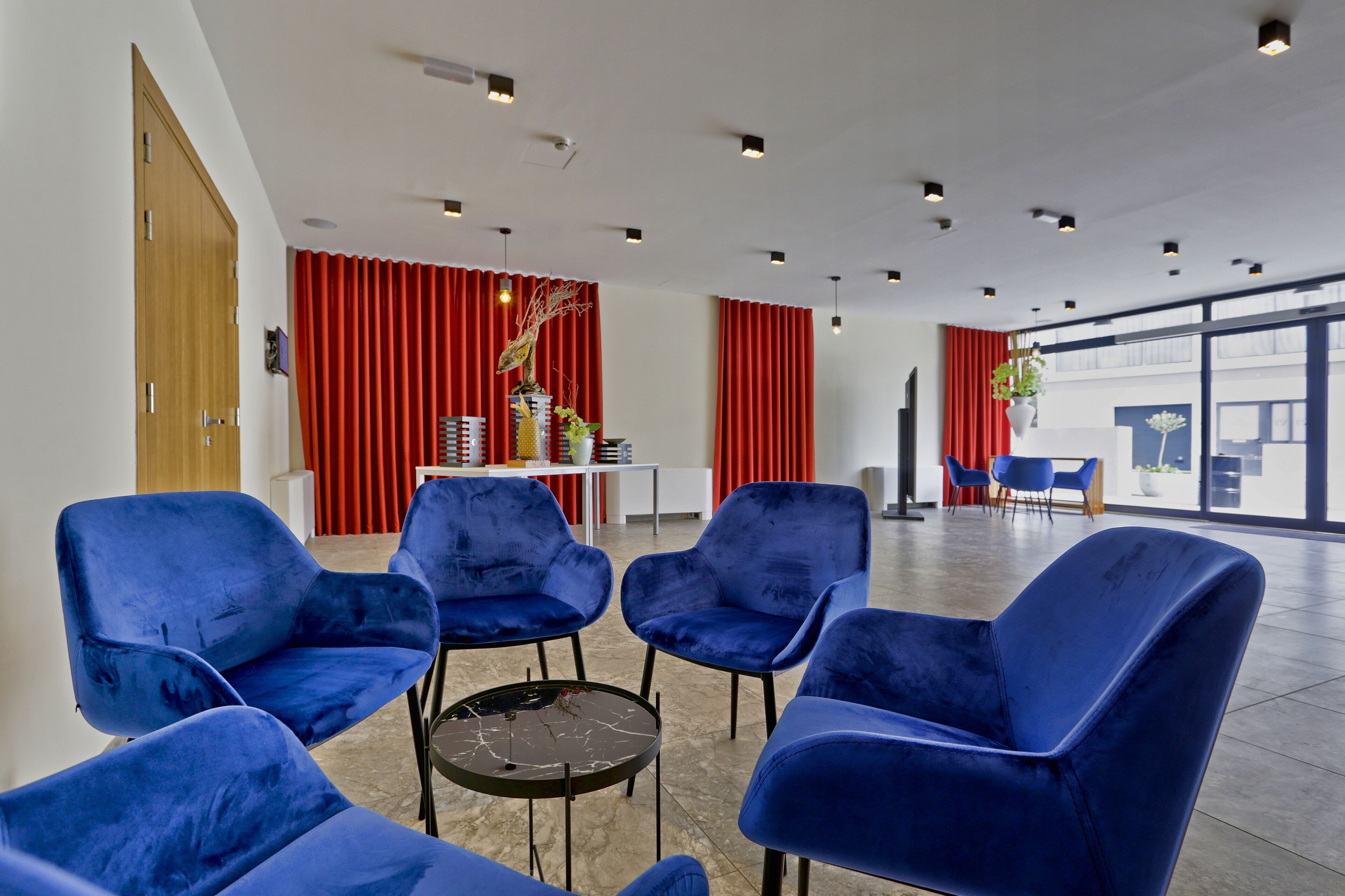 A modern waiting area with blue chairs, a small table, red curtains, and a view through large windows.