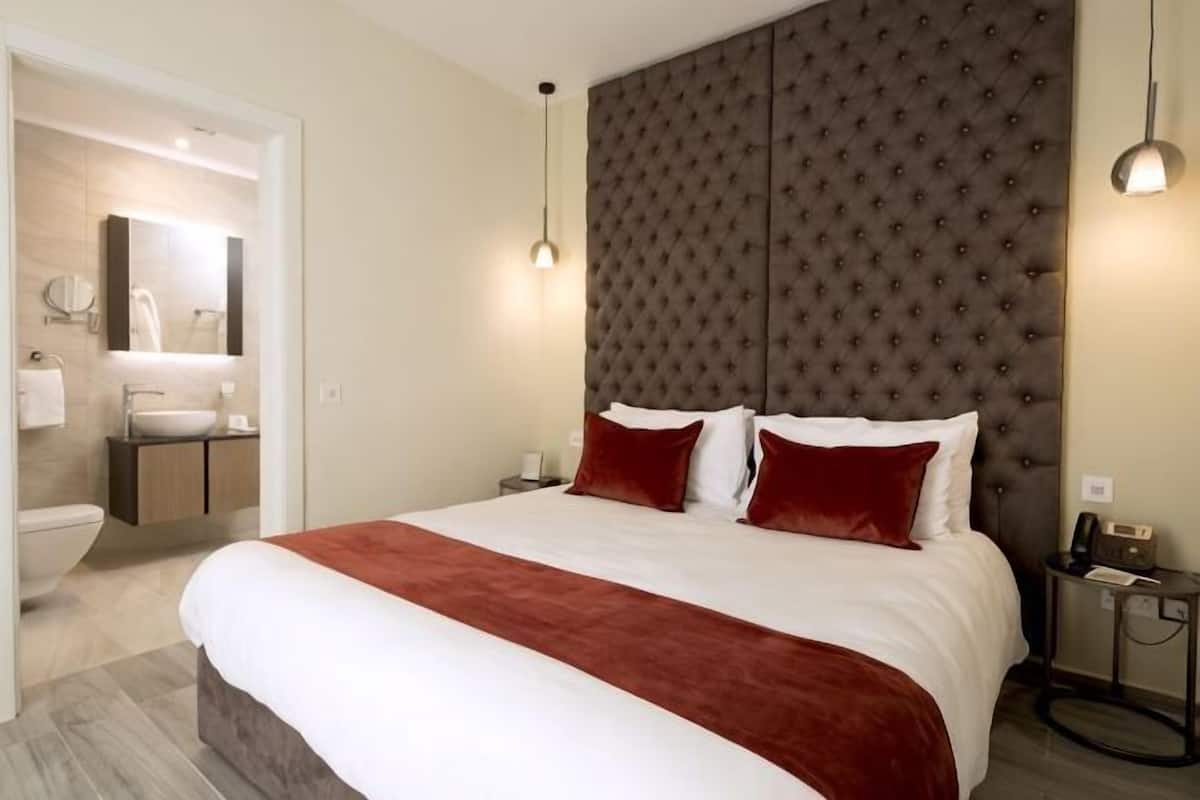 A hotel room with a large bed, a headboard, bedside tables, a nightstand, a mirror, a sink, and a towel rack.