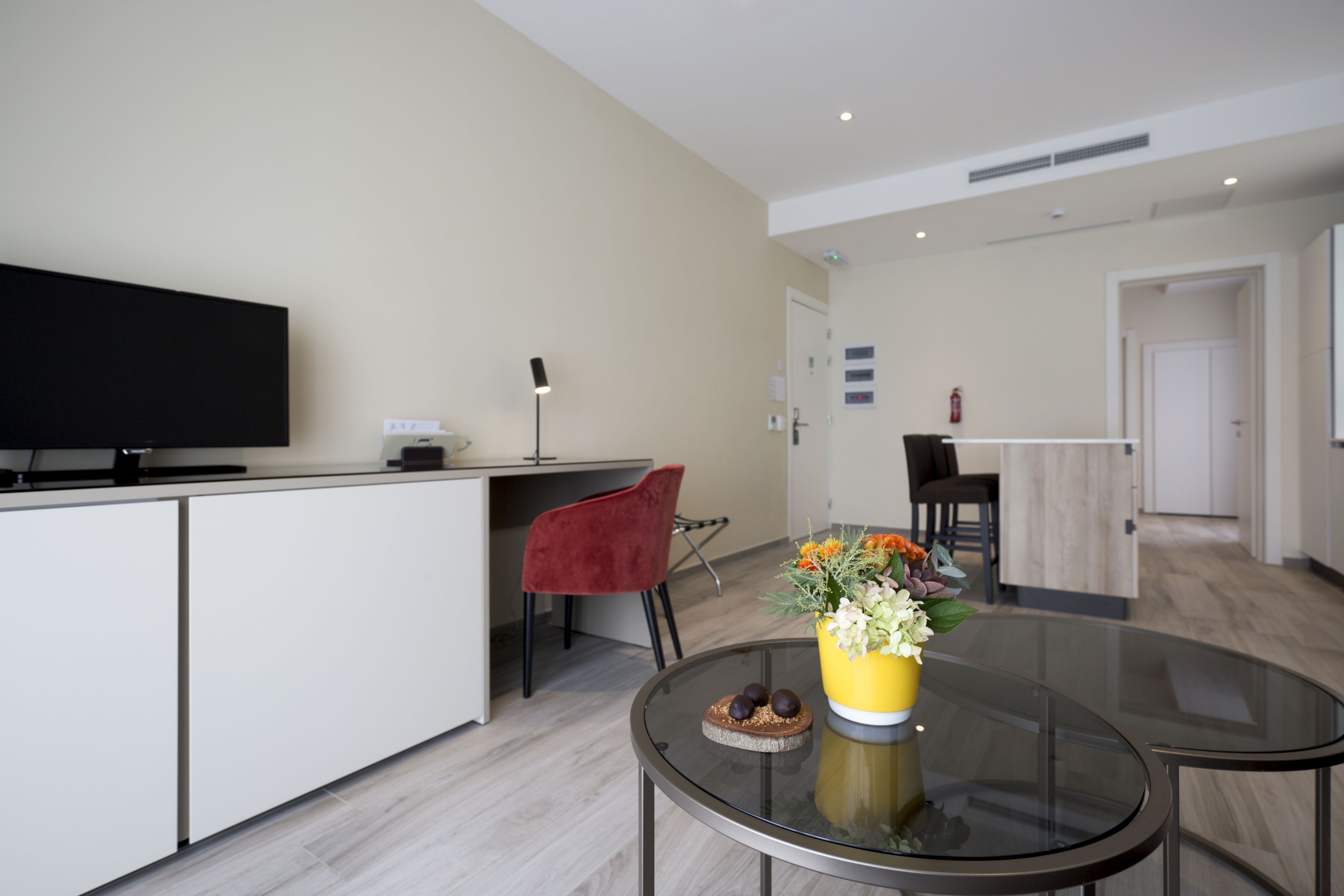 A modern kitchen with a flat-screen TV, a dining area with a glass table, and a red chair.