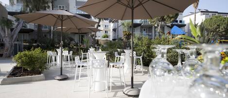 Outdoor seating with umbrellas, white chairs, and tables, surrounded by greenery and buildings.