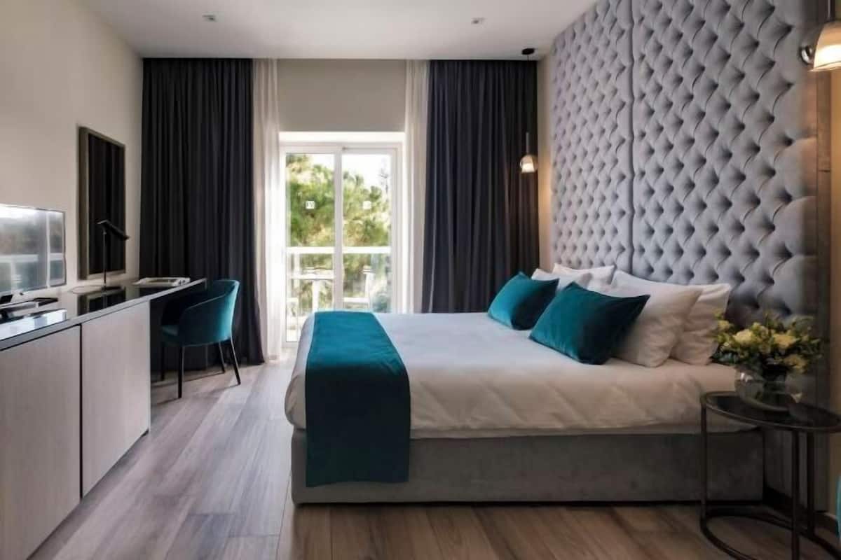 A modern hotel room with a bed, a desk, a chair, and a view of greenery outside.