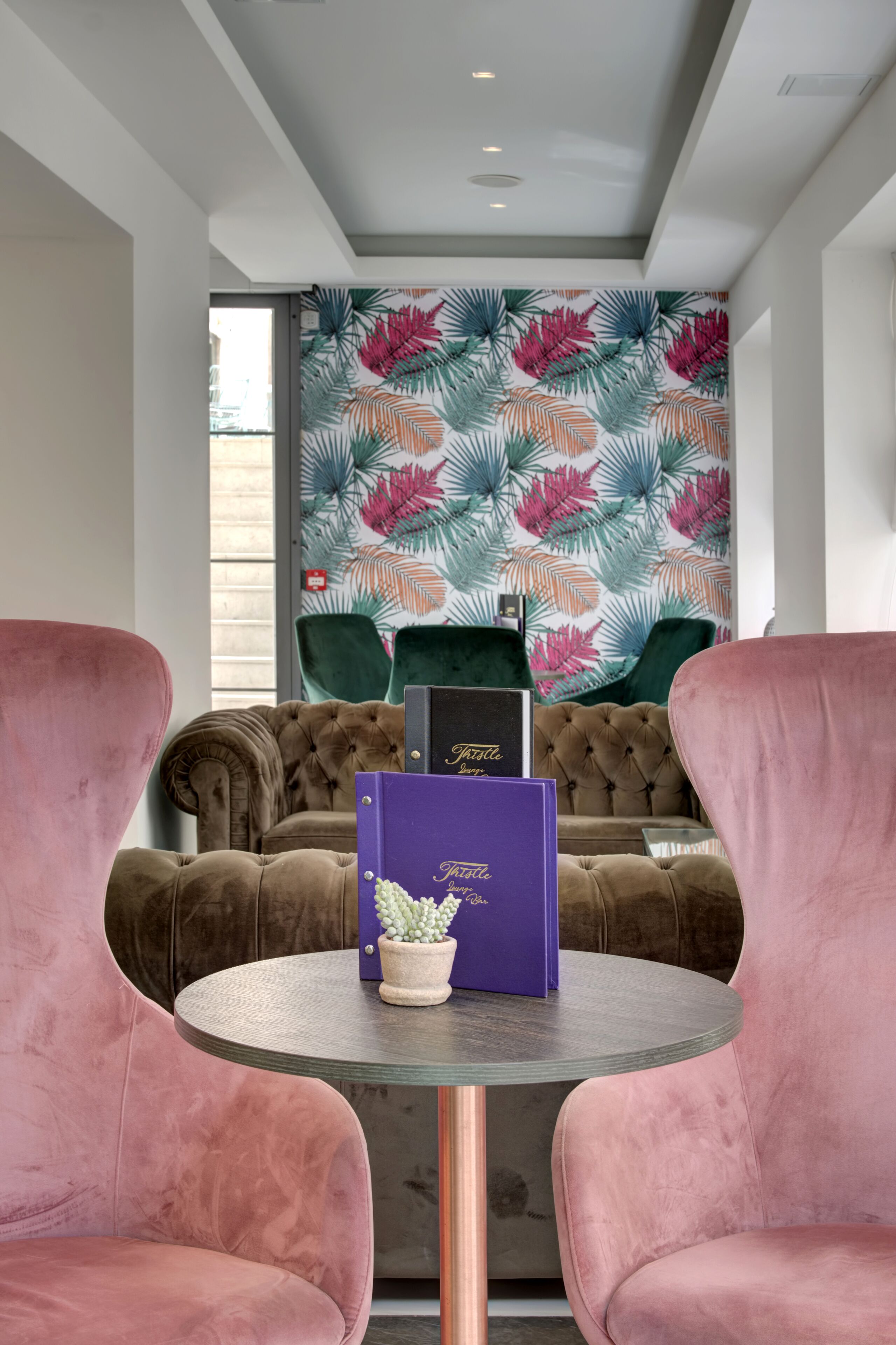 A room with a tufted brown sofa, a round table with a potted plant, and a vibrant tropical leaf wallpaper.