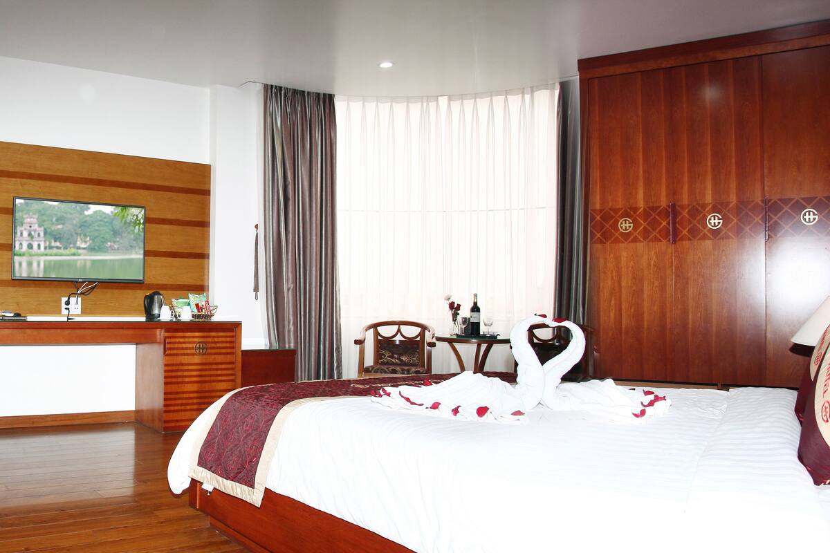 A hotel room with a wooden desk, a television, a bed with a swan-shaped pillow, and a large window with curtains.