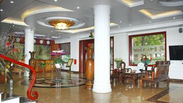 A lobby with a grand staircase, a chandelier, a reception desk, and a seating area with a large window.