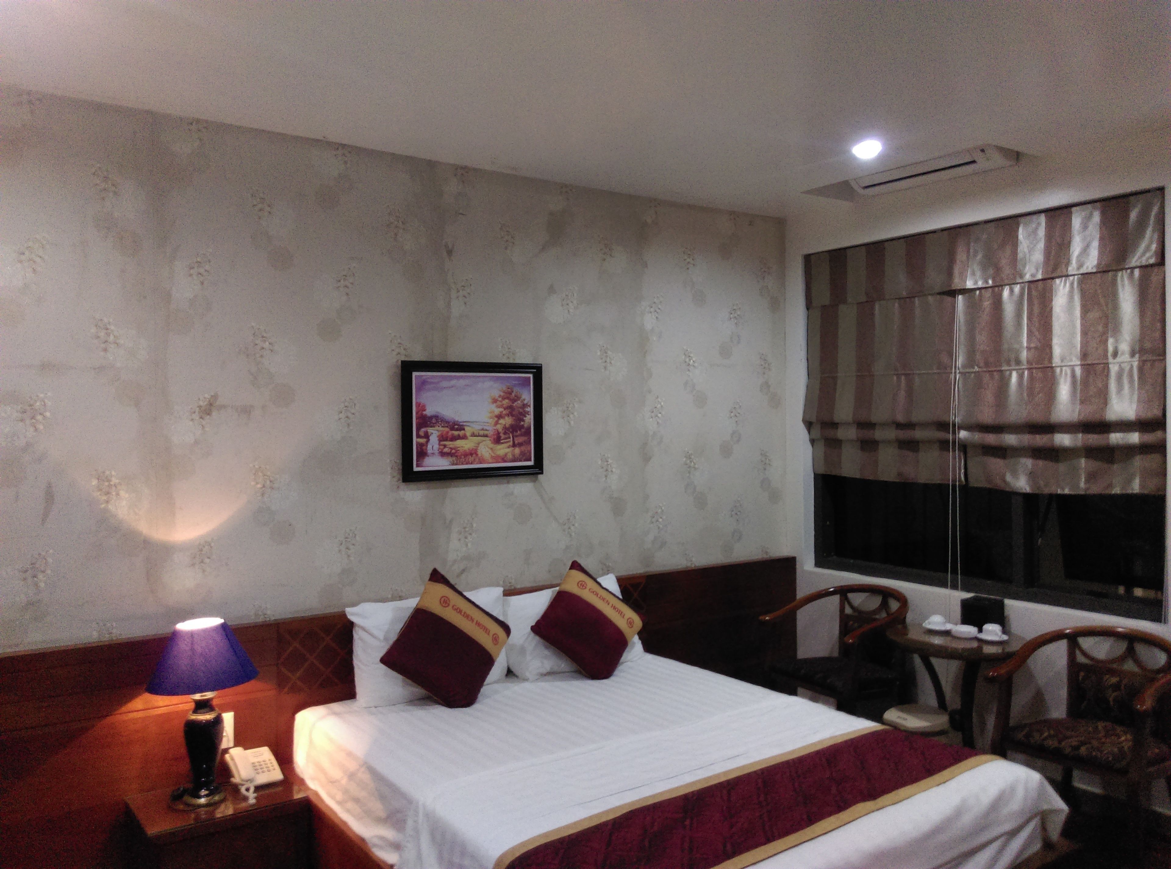 Ein Hotelzimmer mit einem Bett, einem Nachttisch mit Lampe, einem gerahmten Bild an der Wand und einem Fenster mit Vorhängen.