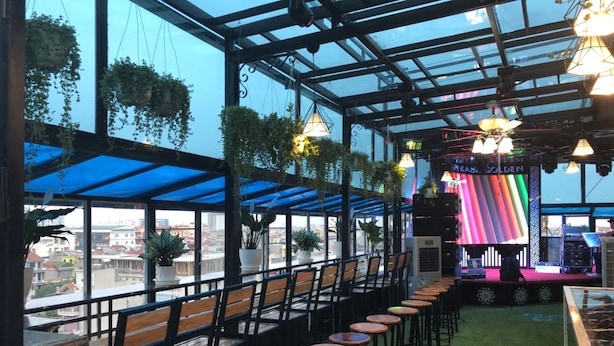 A rooftop bar with rows of wooden bar stools, a stage with colorful lighting, and hanging plants.