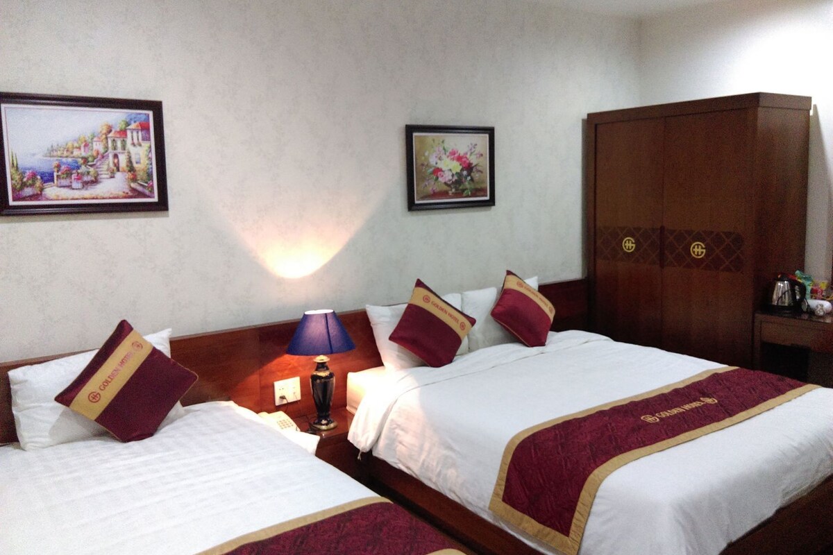 A hotel room with two beds, a nightstand with a lamp, a wardrobe, and two framed pictures on the wall.