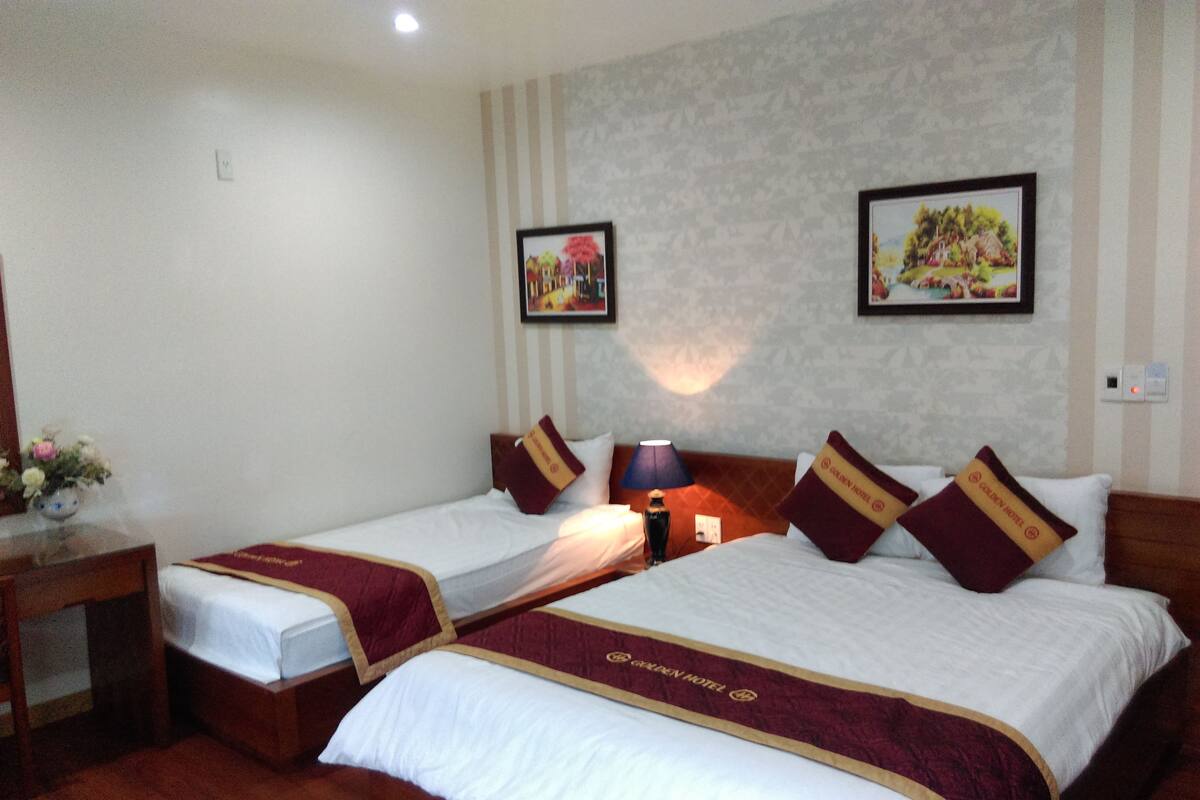 A hotel room with two beds, each with a red and gold bedspread, a nightstand, a lamp, and two framed pictures on the wall.