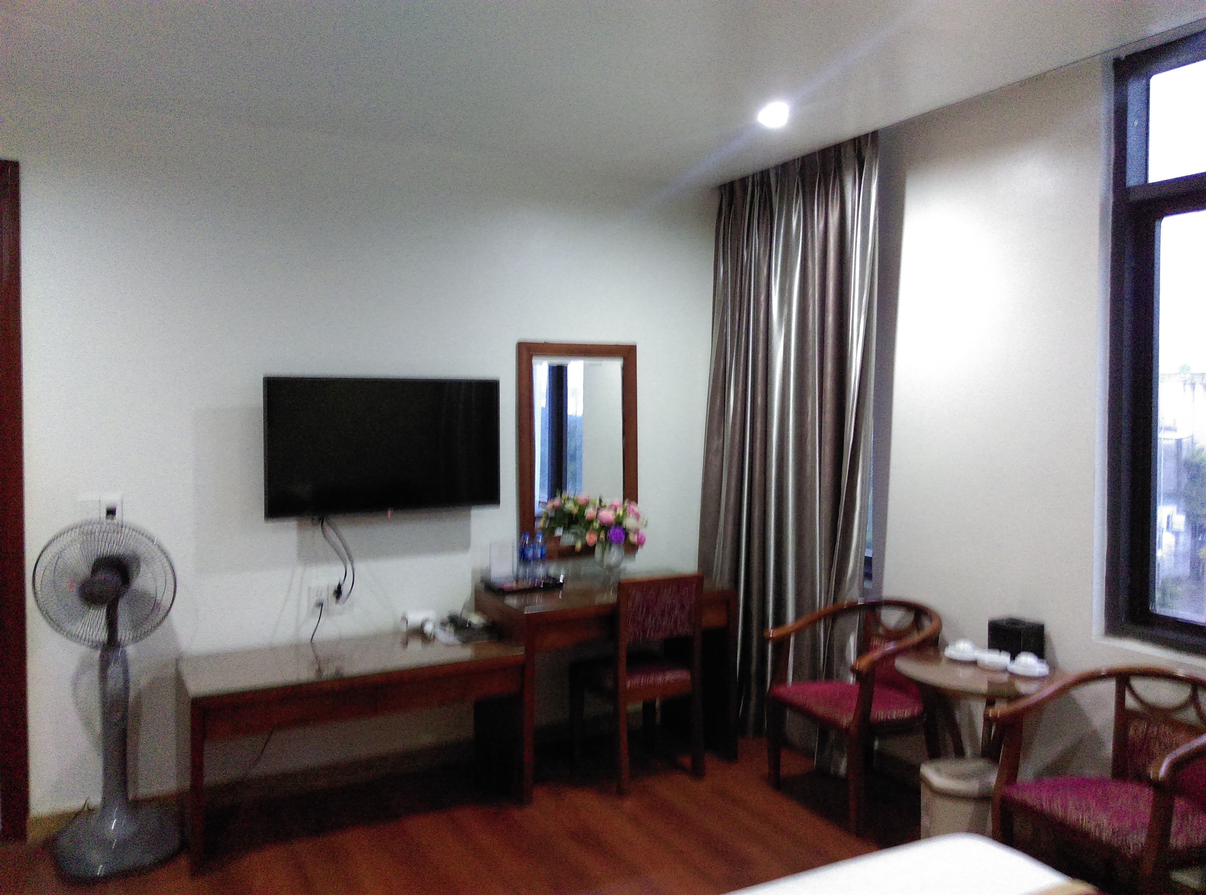 A hotel room with a bed, a desk, a chair, a TV mounted on the wall, a fan, and a window with curtains.