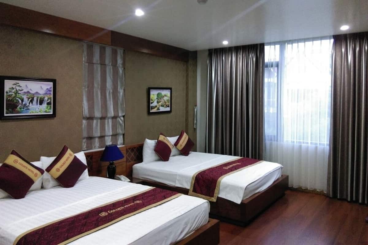 A hotel room with two beds, each with a red and gold headboard, a nightstand with a lamp, and a large window with curtains.
