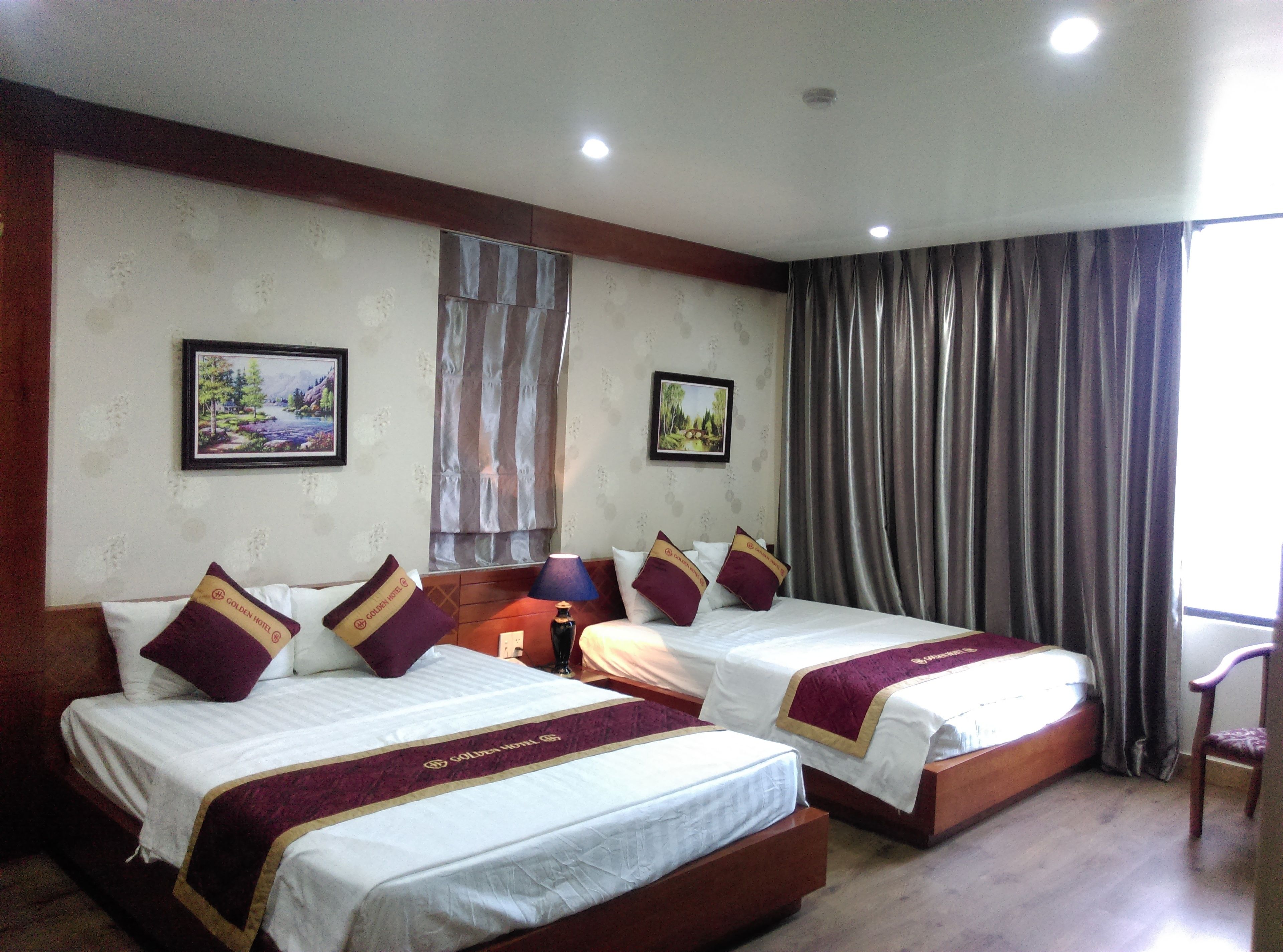 A hotel room with two beds, each with a white mattress and maroon and gold headboards. There are two framed pictures on the wall, a window with curtains, and a wooden chair.