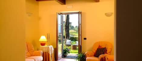 A cozy room with a wooden ceiling, two orange sofas, a small table with a lamp, and a view of a garden through a glass door.
