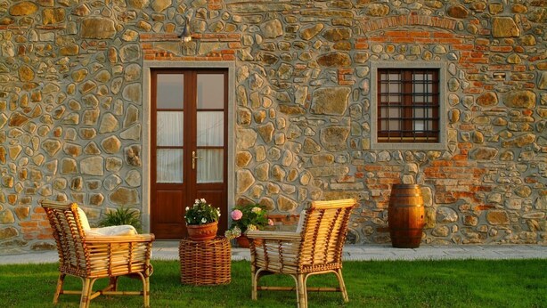Un edificio de piedra con puerta de madera, dos sillas de mimbre, una mesa con flores y una ventana.