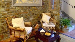 Una habitación con paredes de piedra, dos sillones de mimbre y una mesa de centro de madera, un cuadro abstracto enmarcado y una escalera que lleva a un balcón.