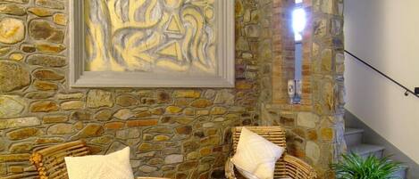 A stone-walled room with two wicker chairs and a wooden coffee table, a framed abstract painting, and a staircase leading to a balcony.