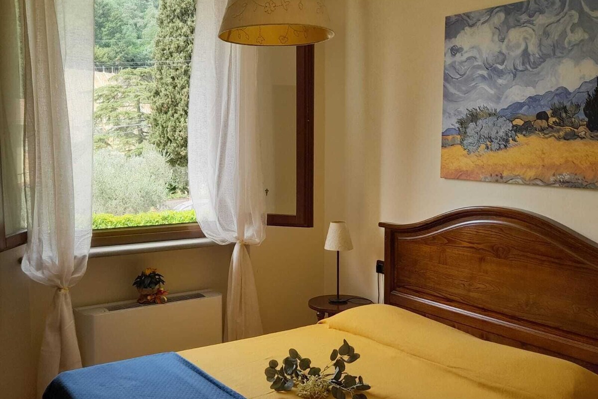 Un dormitorio con cama, ventana con cortinas, mesita de noche con lámpara y un cuadro en la pared.