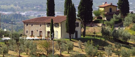A stone building with a red-tiled roof, surrounded by olive trees and cypress trees, set against a hilly landscape.