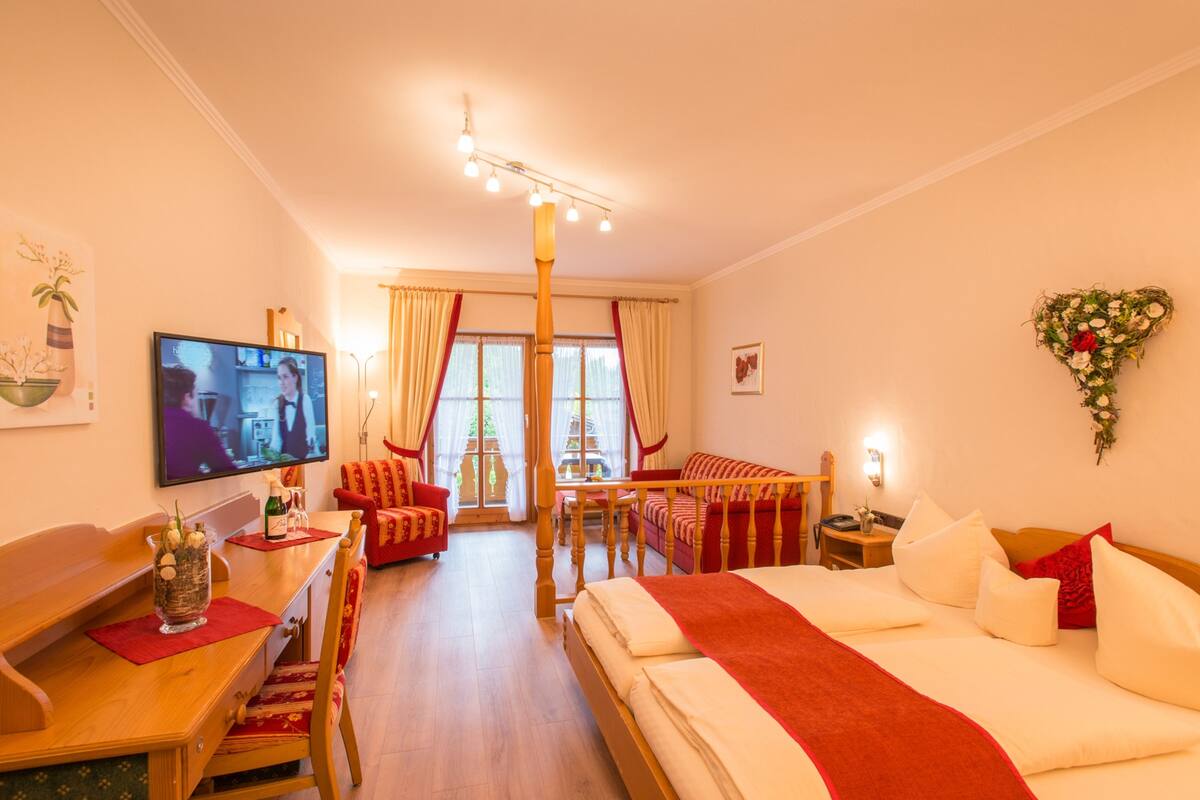 A hotel room with a bed, a TV, a desk with a chair, a dining table with chairs, and a balcony with curtains.
