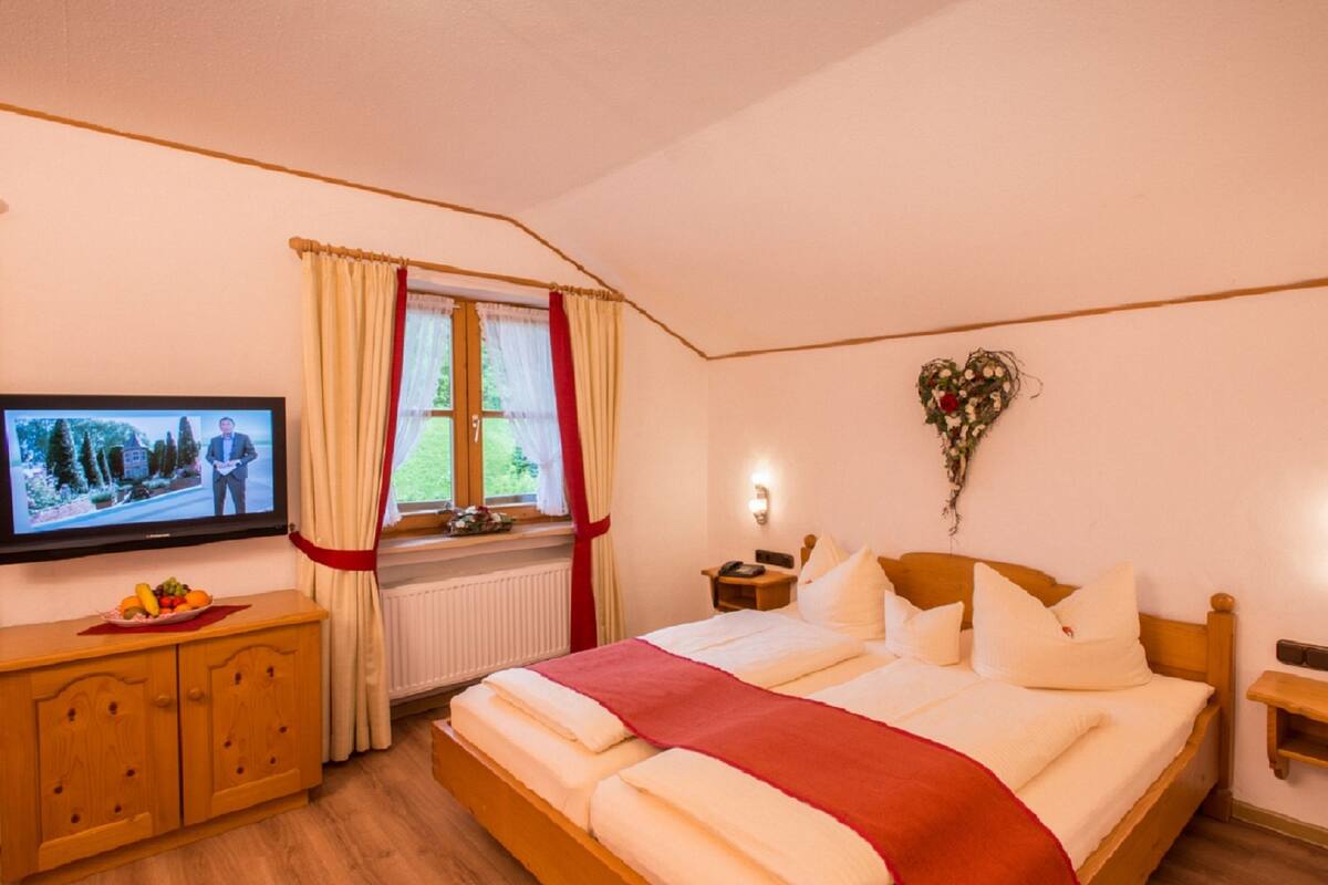 A bedroom with a bed, a television, a window with curtains, a heart-shaped decoration, and a bedside table with a clock.