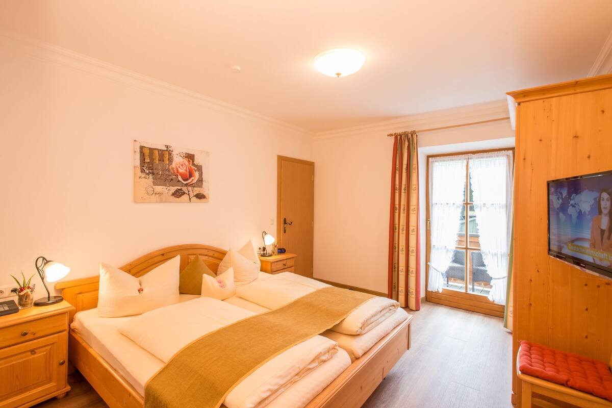 A hotel room with a bed, bedside tables, a TV, a window with curtains, and a painting on the wall.