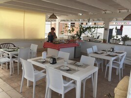 Un restaurante con sillas y mesas de mimbre blanco, una zona de bufé con comida y vistas a edificios al fondo.