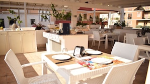 Un área de comedor al aire libre con mobiliario de mimbre blanco, una isla central con cafetera y plantas en macetas.