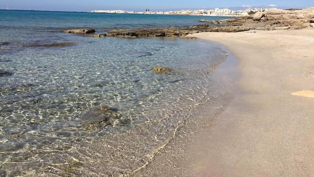 Una playa con aguas cristalinas y poco profundas, una orilla de arena y un horizonte con edificios en la ciudad.