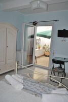Una habitación con una puerta corrediza de vidrio que da a un balcón, un armario blanco, una mesita pequeña y una cama con ropa blanca enrollada.
