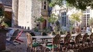 An outdoor dining area with a stone building in the background, a table with chairs, and a tree providing shade. - Chateau d'Agel (Agel)