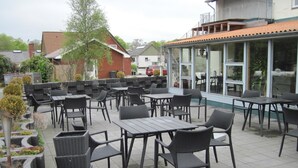 Un patio al aire libre con varias mesas y sillas, rodeado de macetas y un edificio con grandes ventanales.